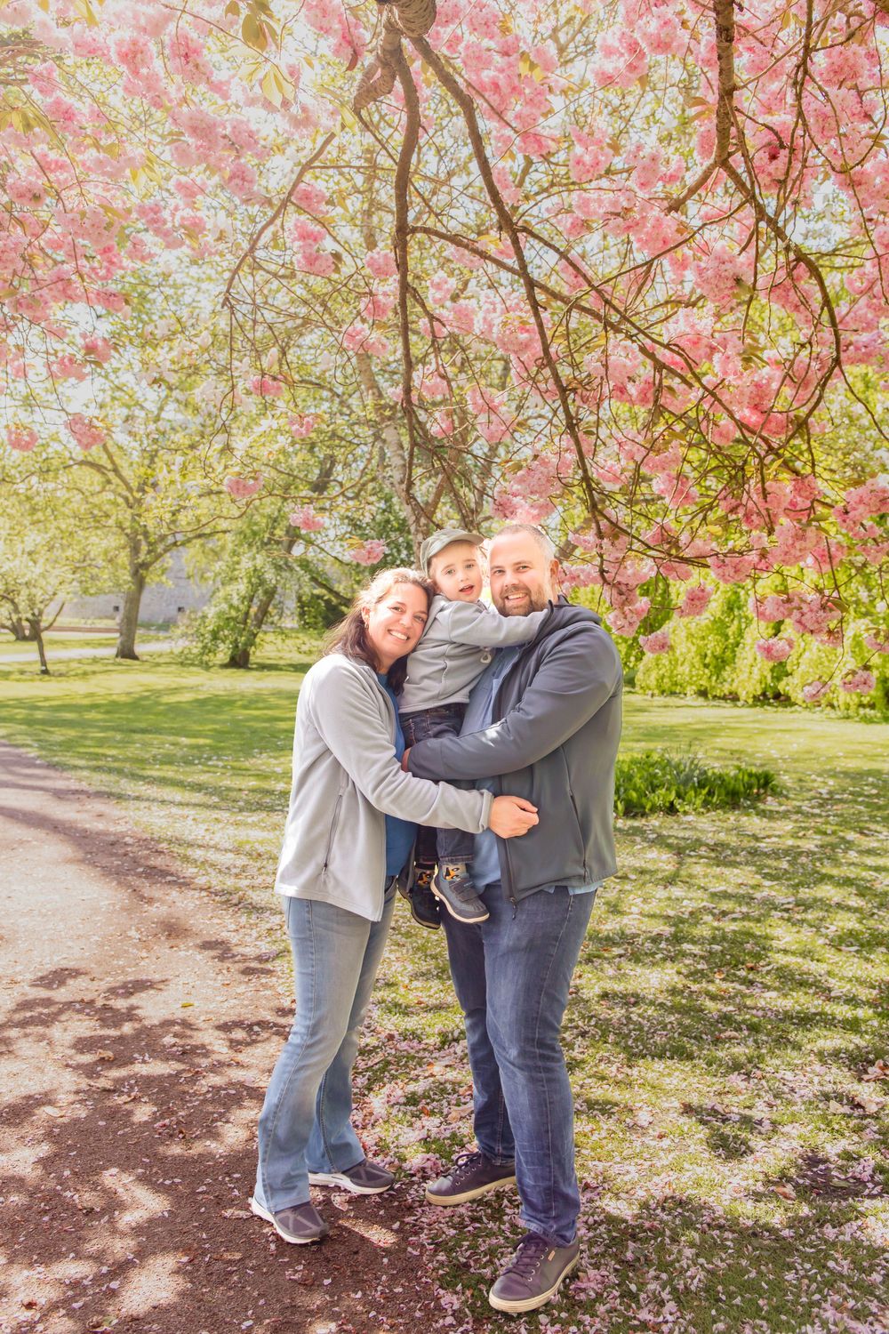 Happy family of three hugging under pink cherry blossom trees in a sunny park during spring.