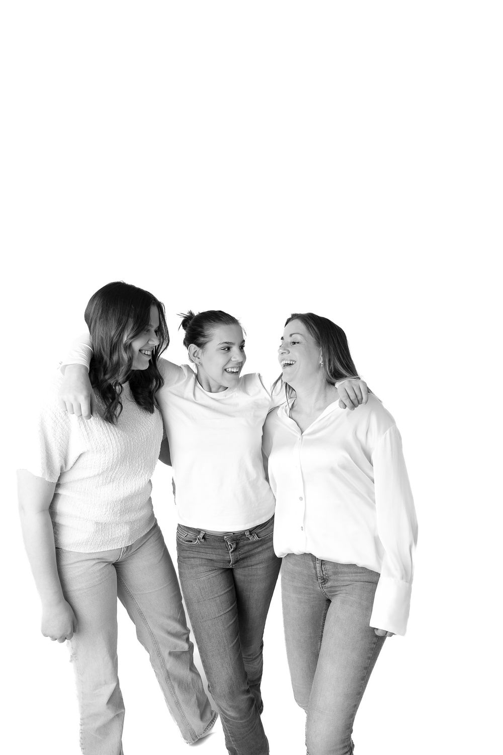 Three women smiling and embracing in a black and white studio photo, wearing casual tops and jeans.