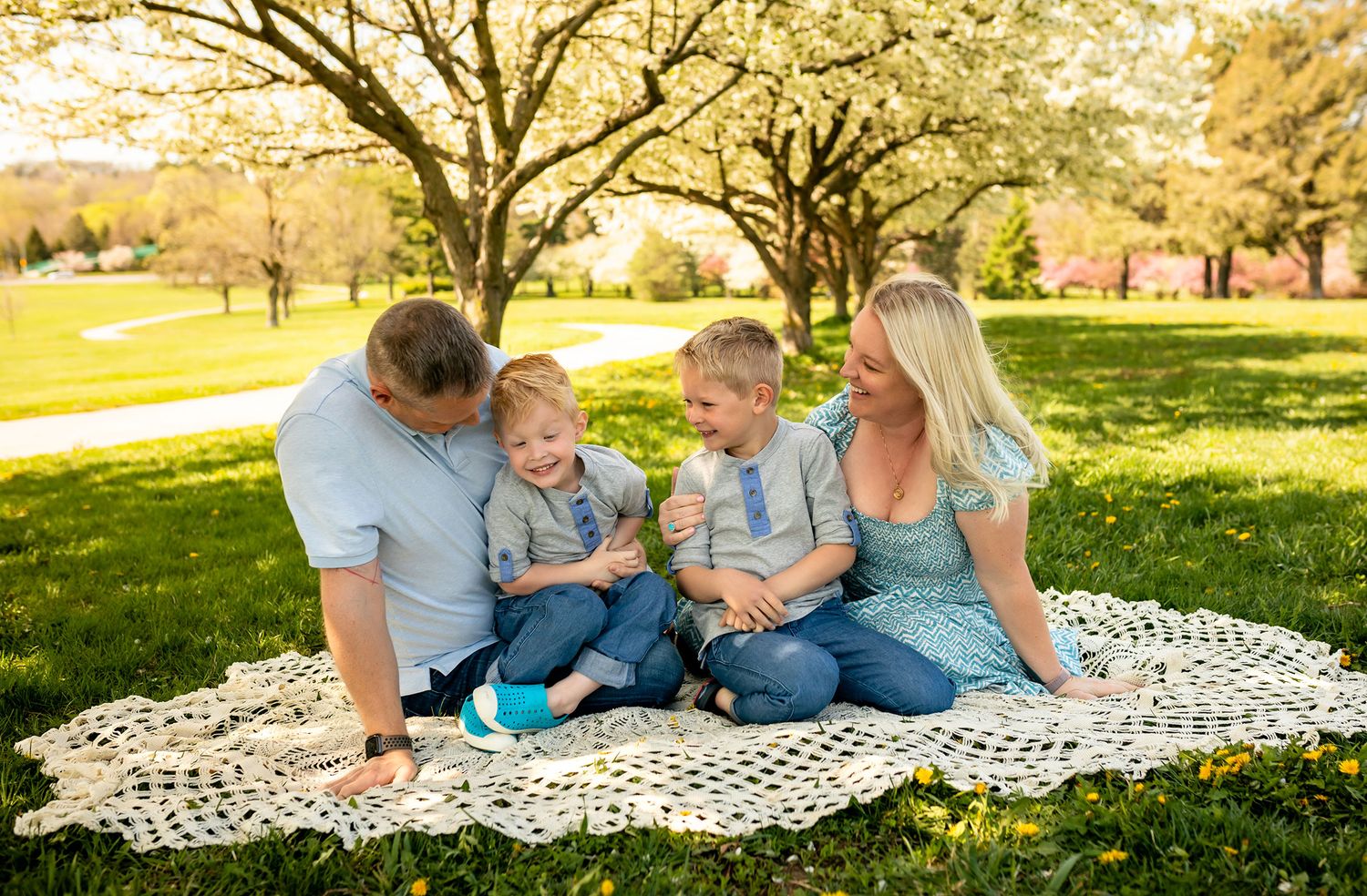 Family of four sitting on blanket under blooming trees in sunny park, parents smiling at two young blonde boys.