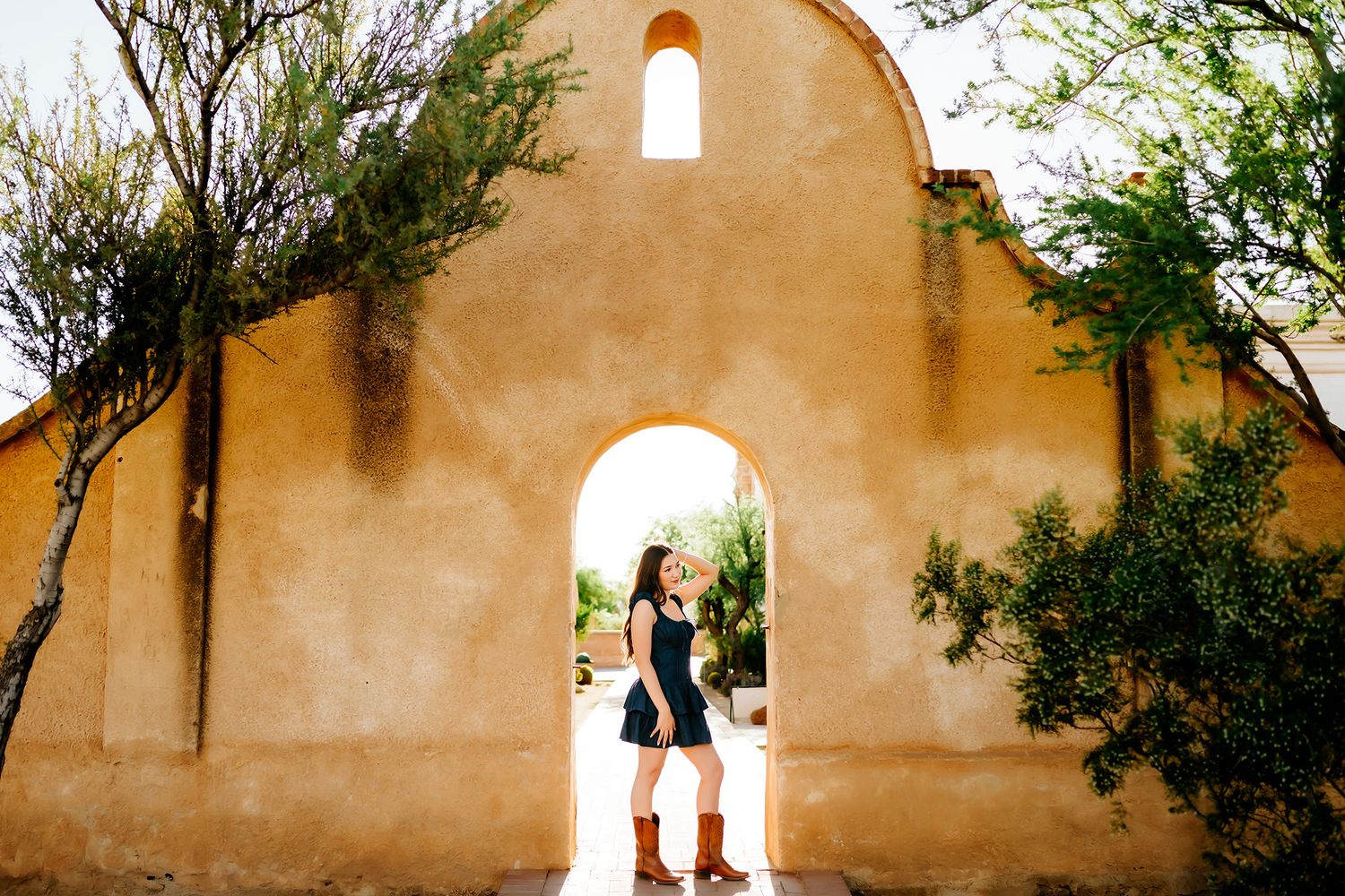 Tucson senior photographer capturing high school senior girl posing in adobe archway with warm golden light, blue diem dress and boots