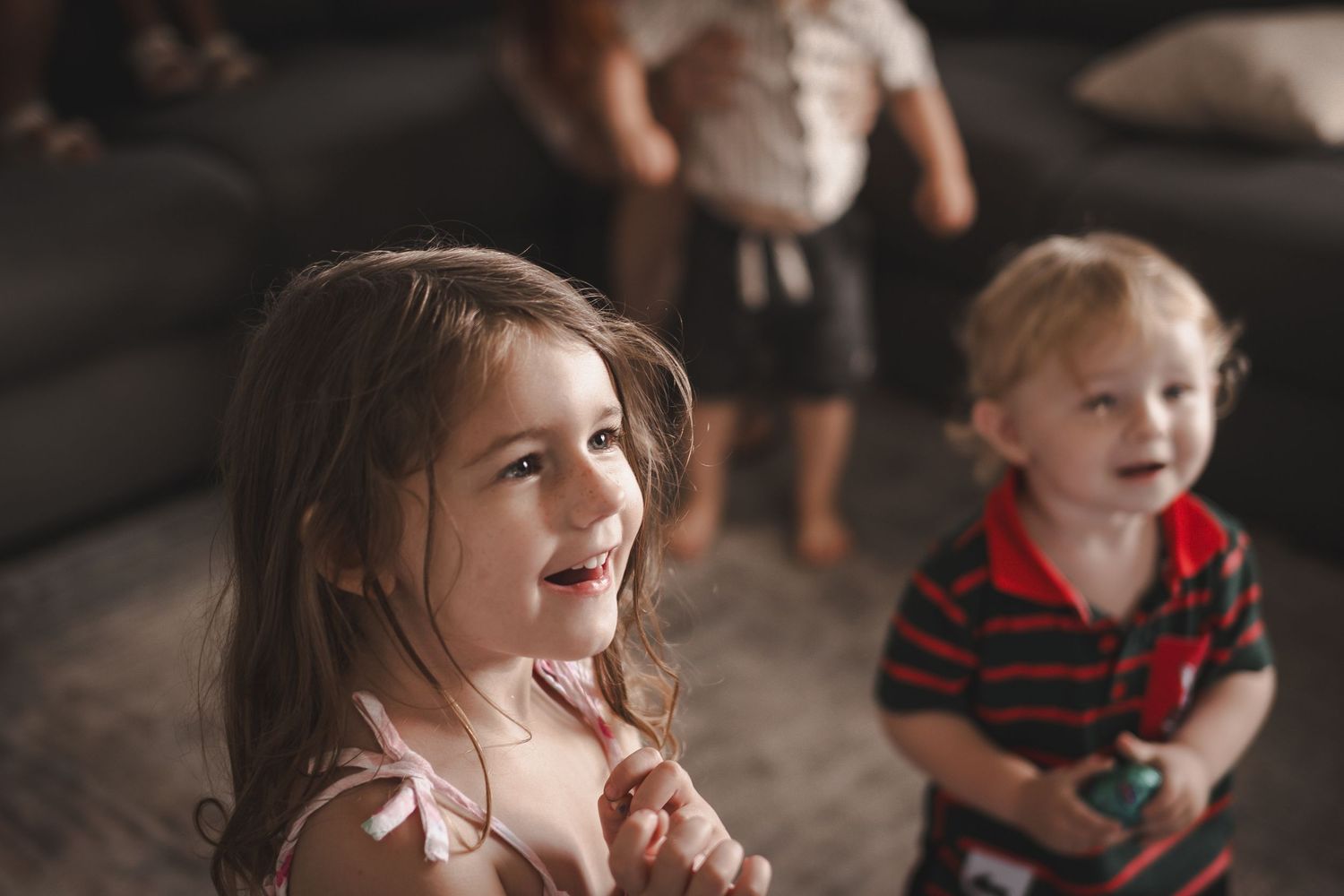 A young girl smiles with hands clasped while a toddler boy sits nearby in a cozy living room.