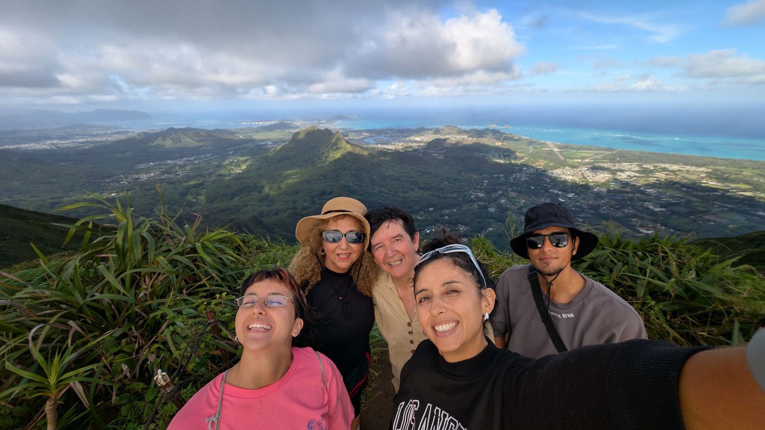 Five friends take a selfie at a scenic mountain overlook in Hawaii with lush green hills and ocean views behind them.