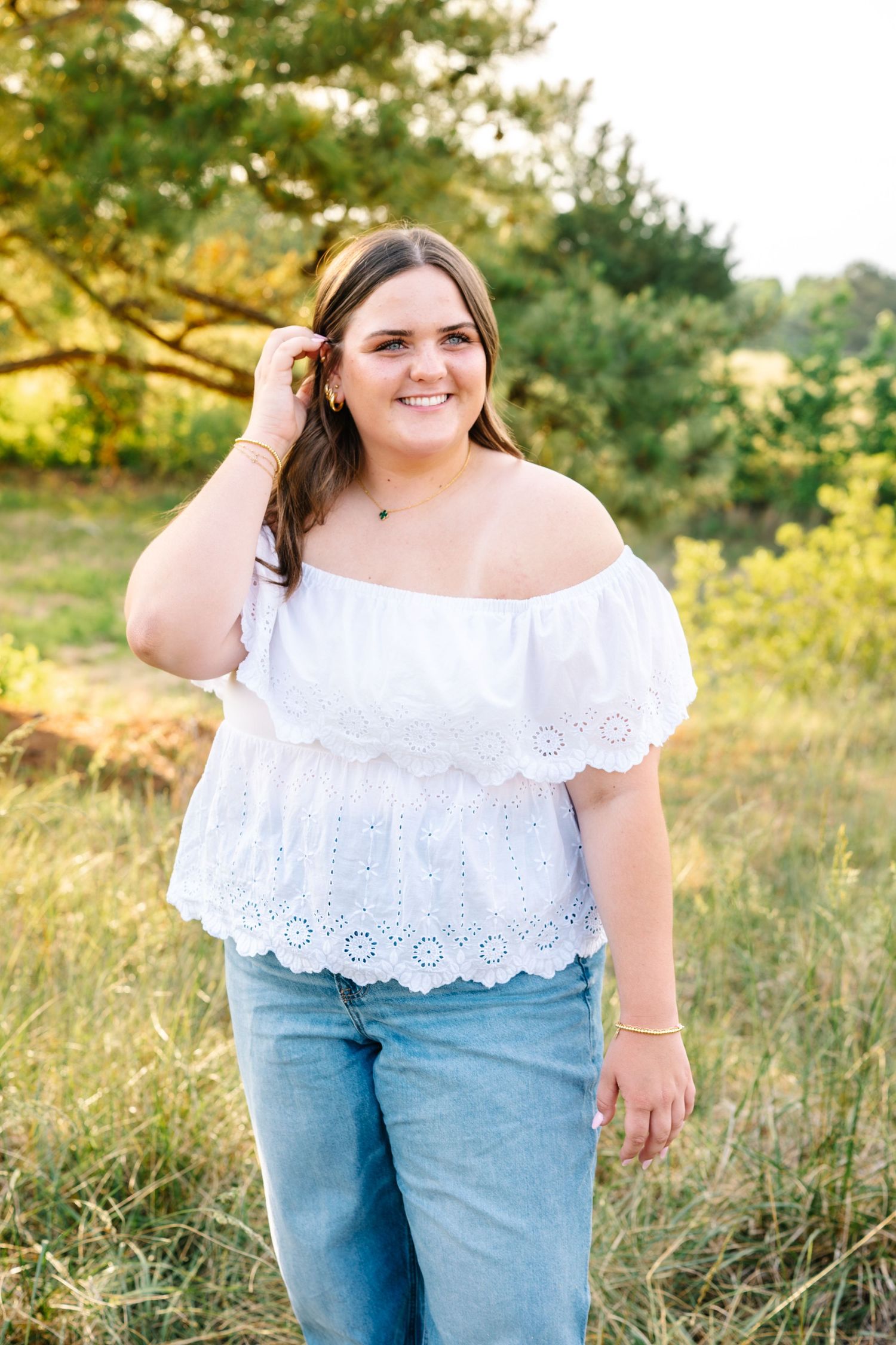Young woman in white off-shoulder eyelet top and jeans smiling outdoors in a sunny field.