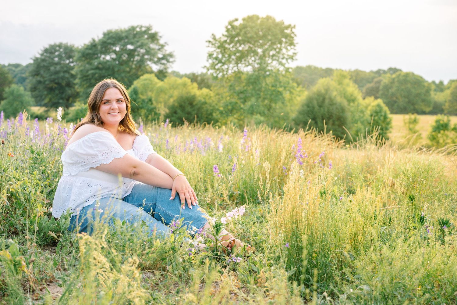 A smiling woman sits in a lush wildflower meadow wearing a white top and jeans during golden hour.