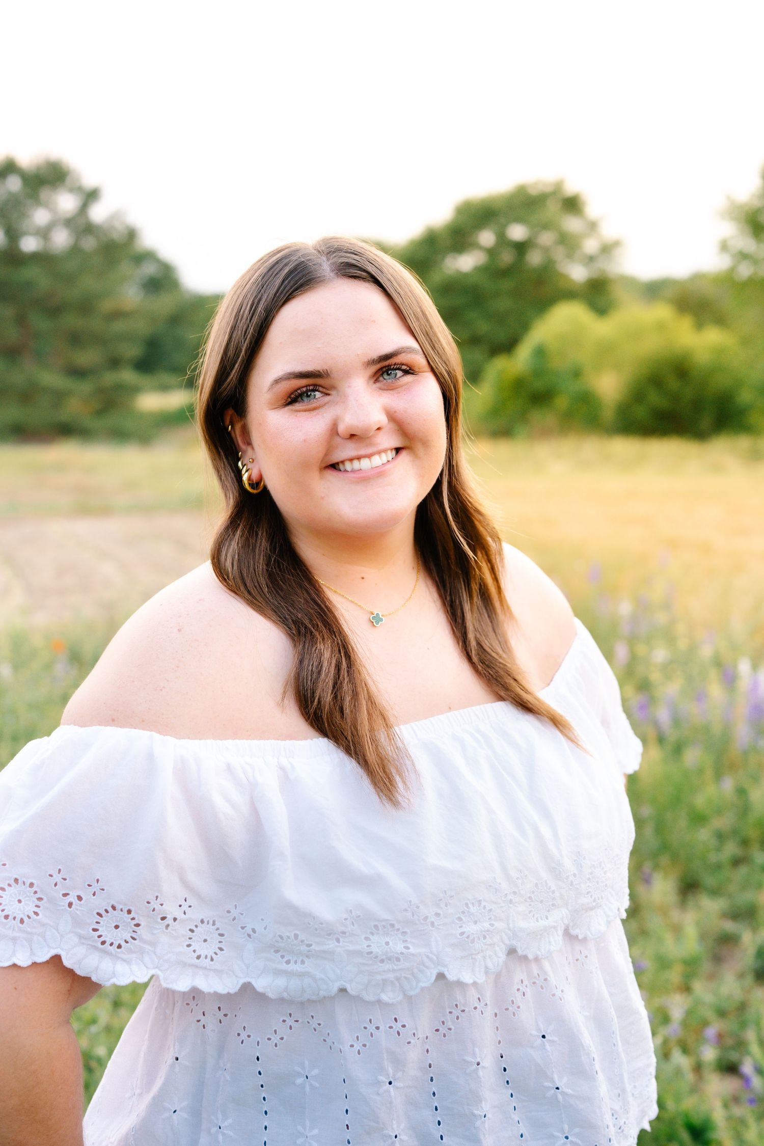 Young woman in white off-shoulder dress smiling in a sunny field with green trees in the background.