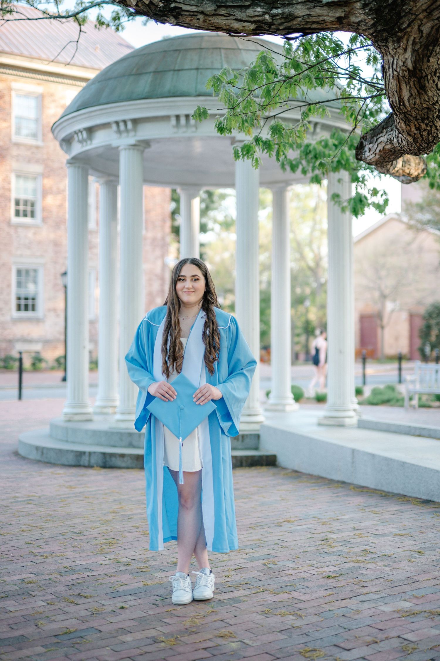 Young woman in light blue graduation cap and gown standing near white rotunda columns on a college campus.