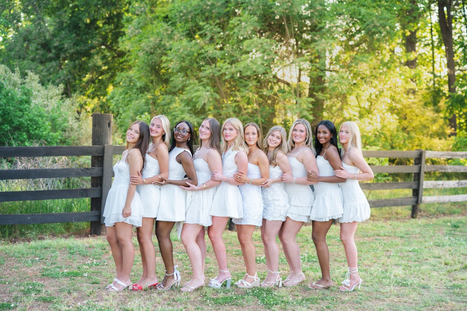 Group of nine young women in white dresses pose together smiling outdoors near wooden fence.