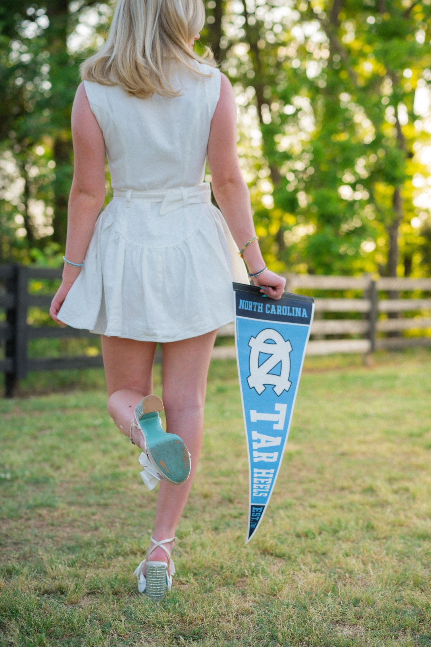 Blonde girl walks away holding North Carolina Tar Heels pennant in white dress and green socks.