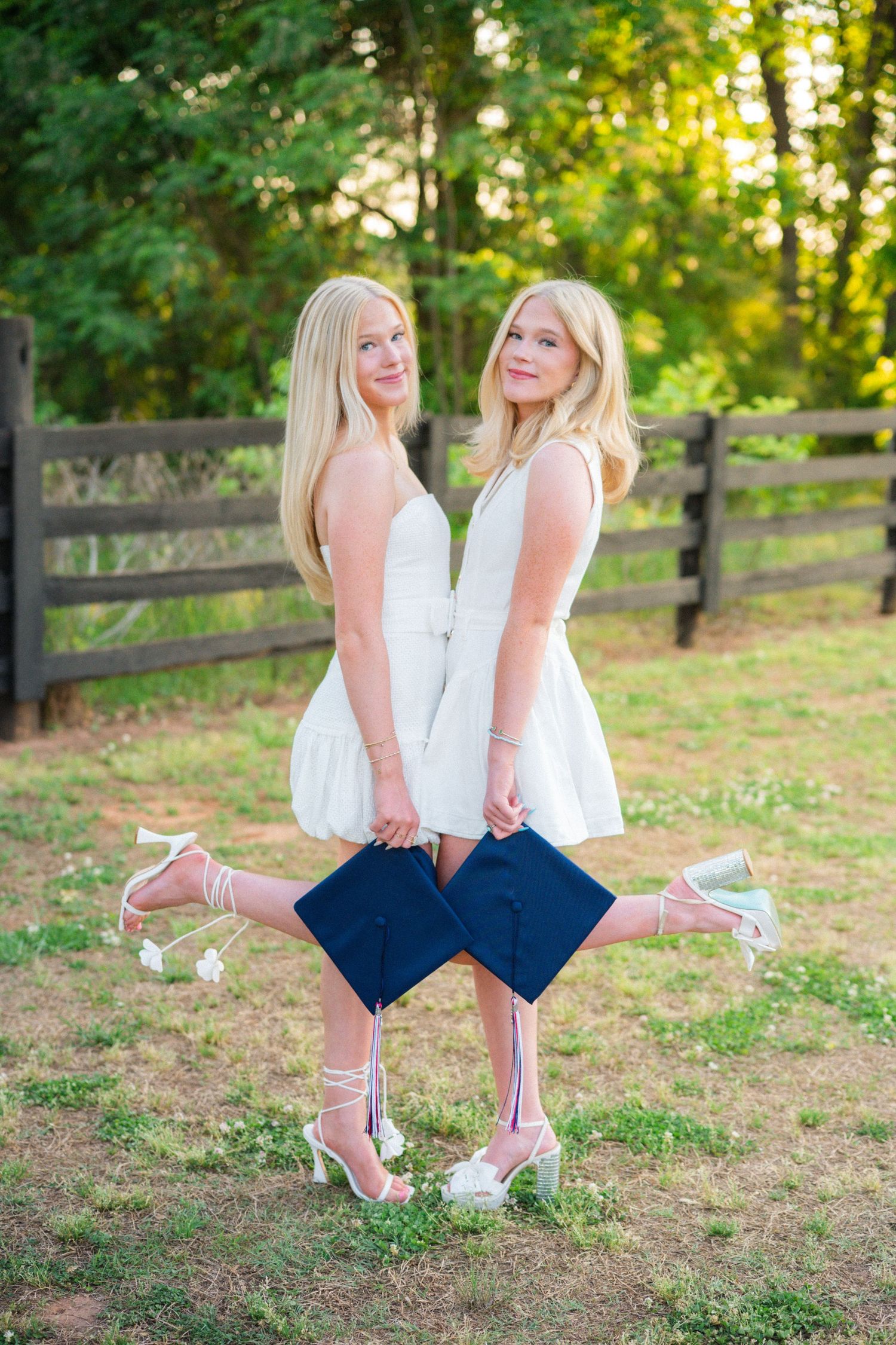 Two blonde twin girls in white dresses holding navy graduation caps posing back-to-back outdoors at golden hour.
