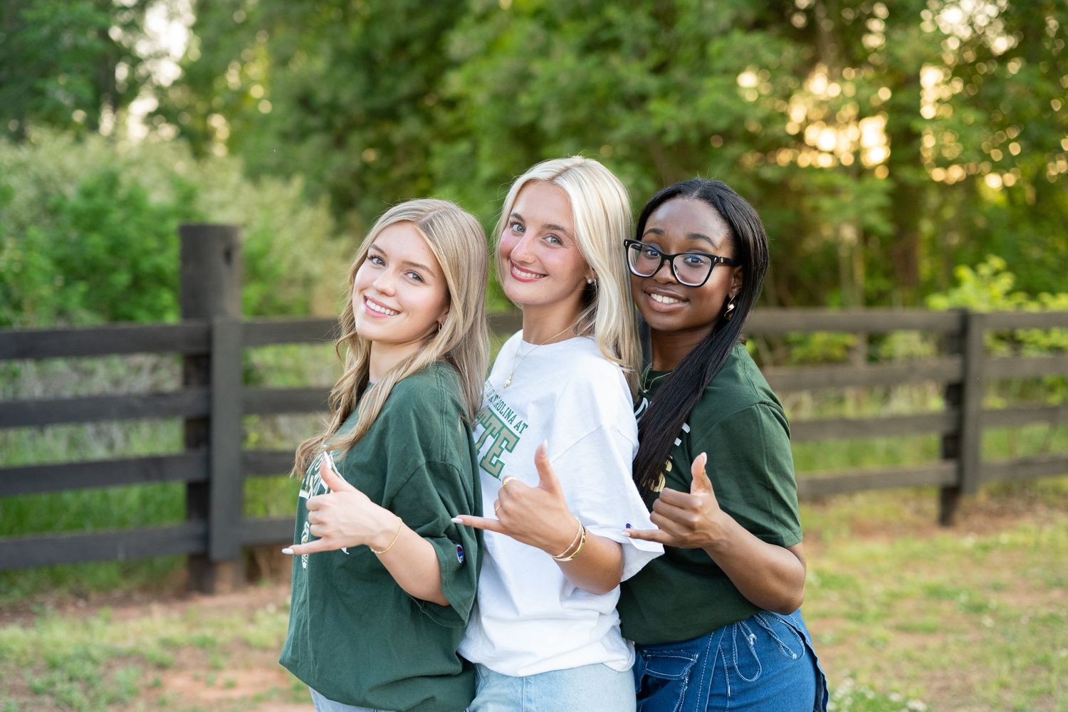 Three smiling young women pose outdoors in green and white shirts near a wooden fence.