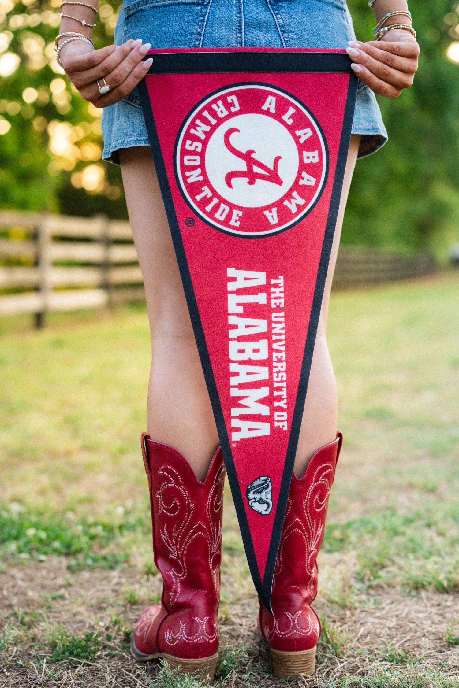 University of Alabama Crimson Tide pennant held by person wearing red cowboy boots outdoors.