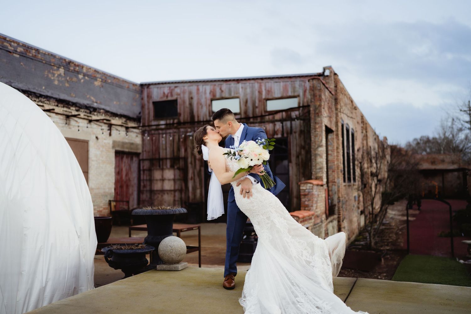 Bride and groom share romantic dip kiss outdoors at rustic wedding venue with historic brick building backdrop.