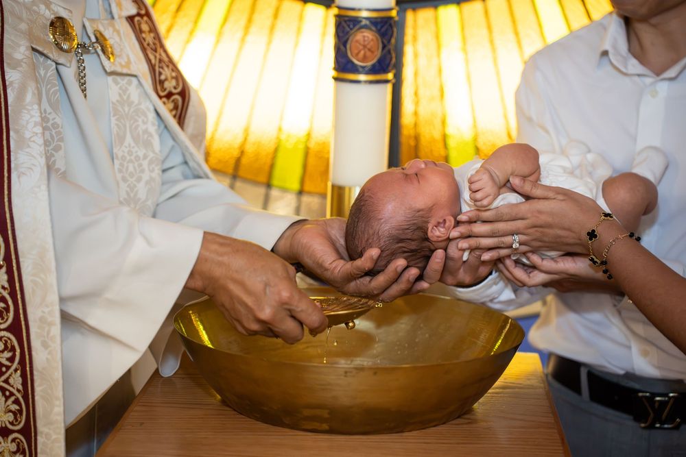 Father and godmother supporting an infant as the priest performs a baptism at Holy Spirit in Tempe, by N&D Media Production.