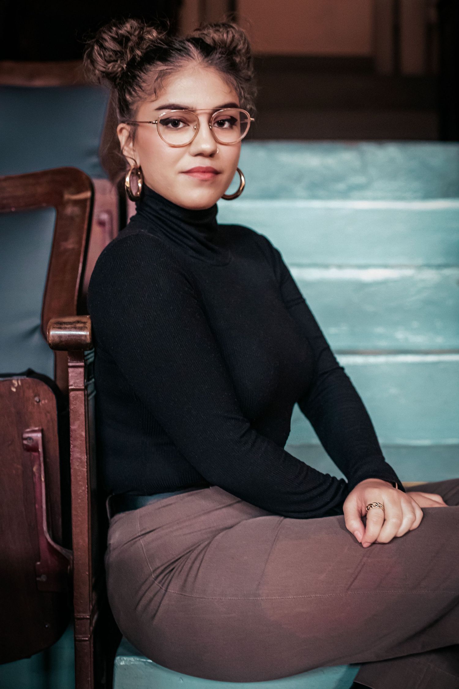 A young woman with glasses and curly hair is sitting on a chair and looking at the camera.