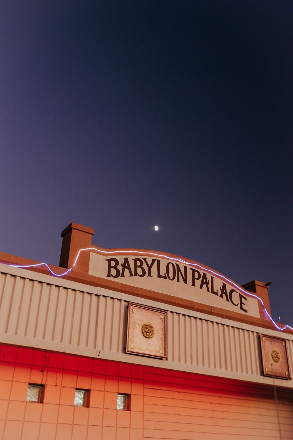 A vintage-style sign reading 'Babylon Palace' on a beige building exterior with a moon visible in the dark sky.