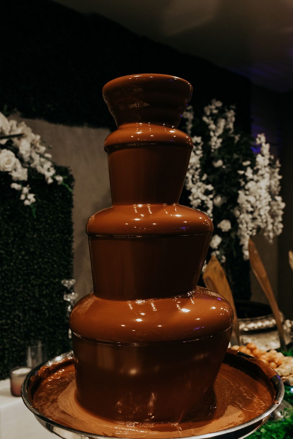 Three-tiered chocolate fountain with flowing melted chocolate surrounded by white floral decorations.