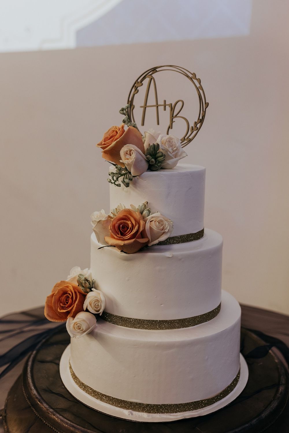 Elegant white wedding cake with gold accents and peach roses displayed on a dark stand.
