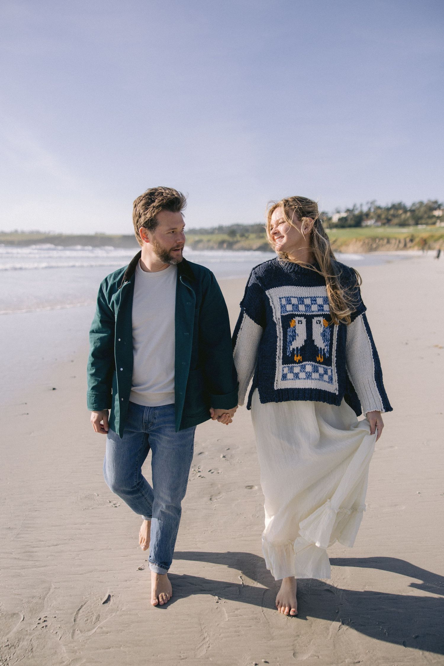 A couple walks barefoot on a sandy beach, wearing casual winter clothing and holding hands on an overcast day.