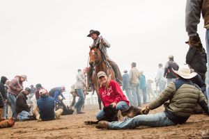 Migrants rest on dusty ground while border patrol officers on horseback patrol the perimeter in foggy conditions.