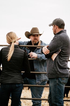 Three people having a conversation while leaning on wooden fence rails at a ranch.