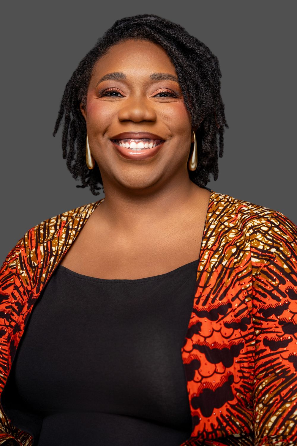 A person wearing a black top and vibrant orange patterned jacket poses against a gray background with a bright smile.
