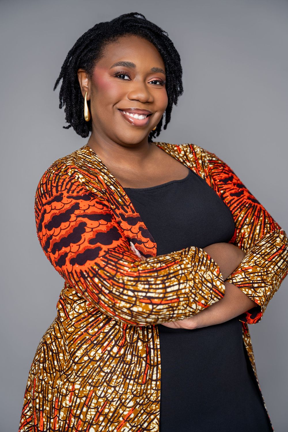 A person wearing a black dress with vibrant orange and gold African print puff sleeves poses against a gray background.
