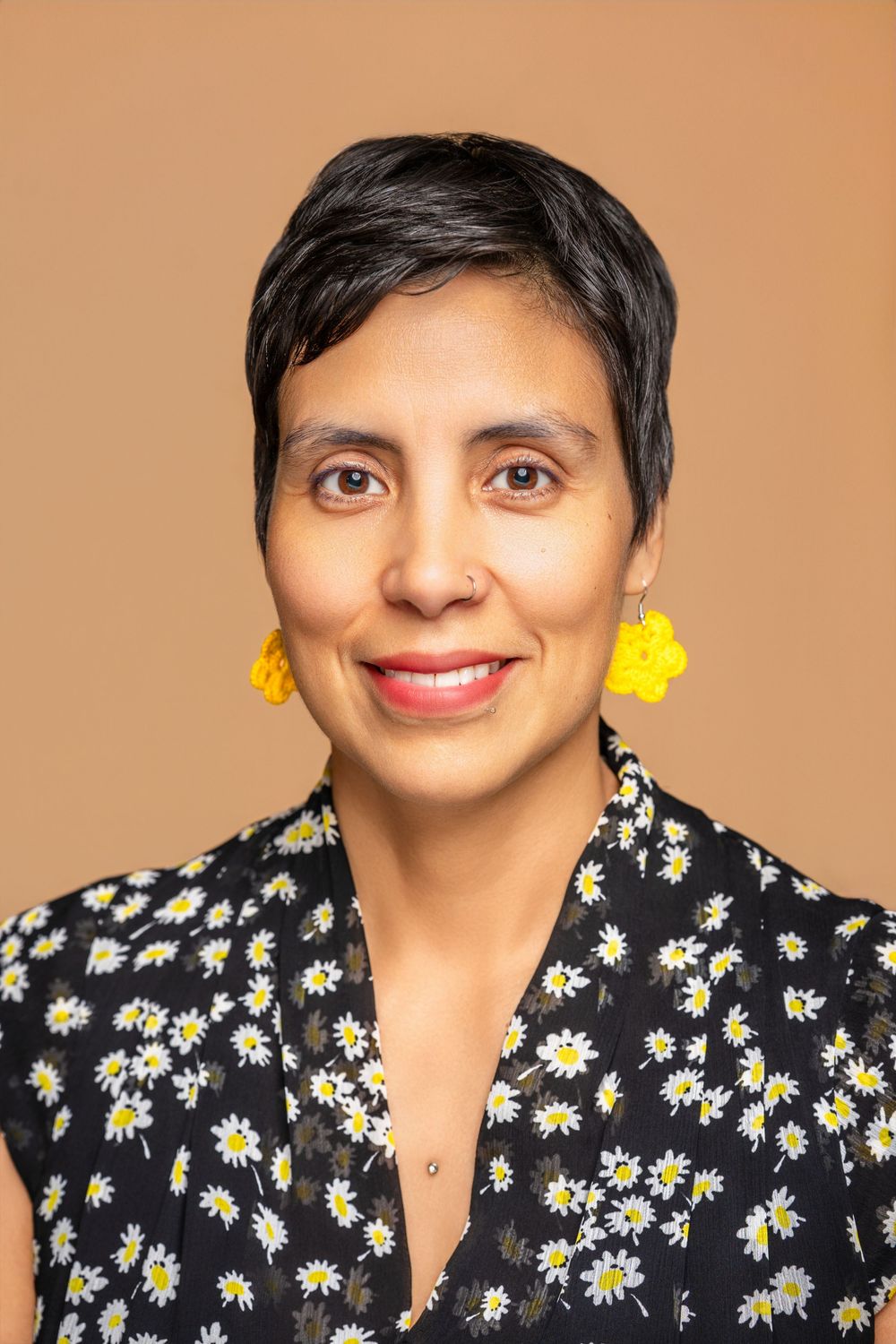 Woman with short black hair wearing yellow floral earrings and daisy print blouse, smiling against beige background.