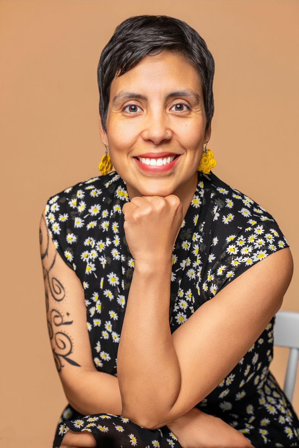 Smiling woman with short hair, daisy-print dress, yellow tassel earrings, and arm tattoo poses on beige background.