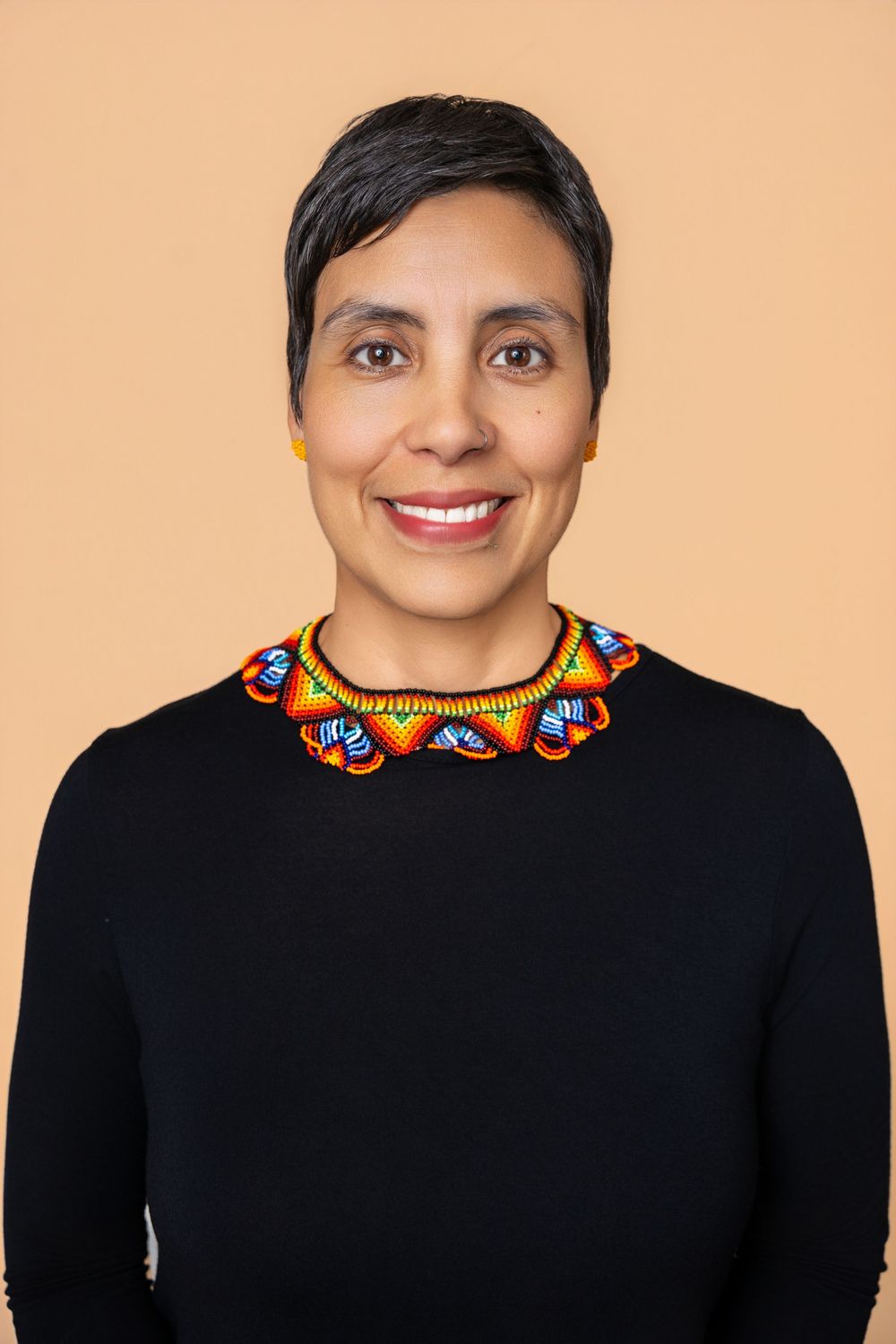 Smiling woman with short hair wearing a black top and colorful beaded necklace against a beige background.