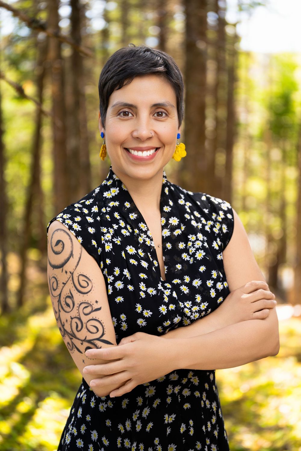 Smiling woman with short hair, floral dress, tattoo, and colorful earrings posing outdoors in a forest setting.