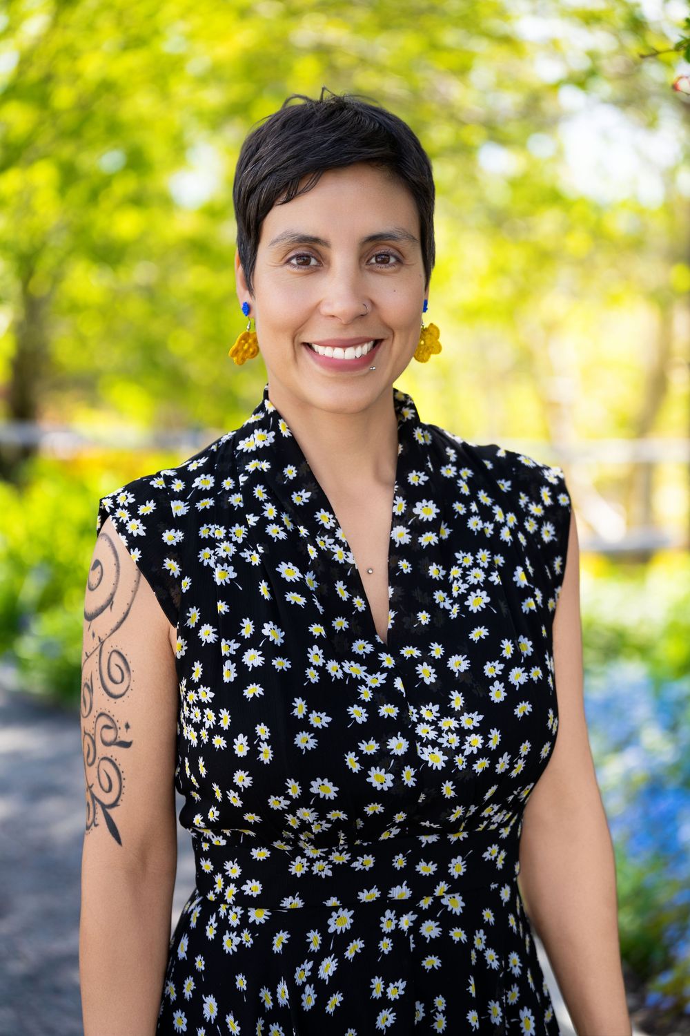 Woman in floral dress with tattoo and yellow earrings smiling outdoors in a lush green garden setting.