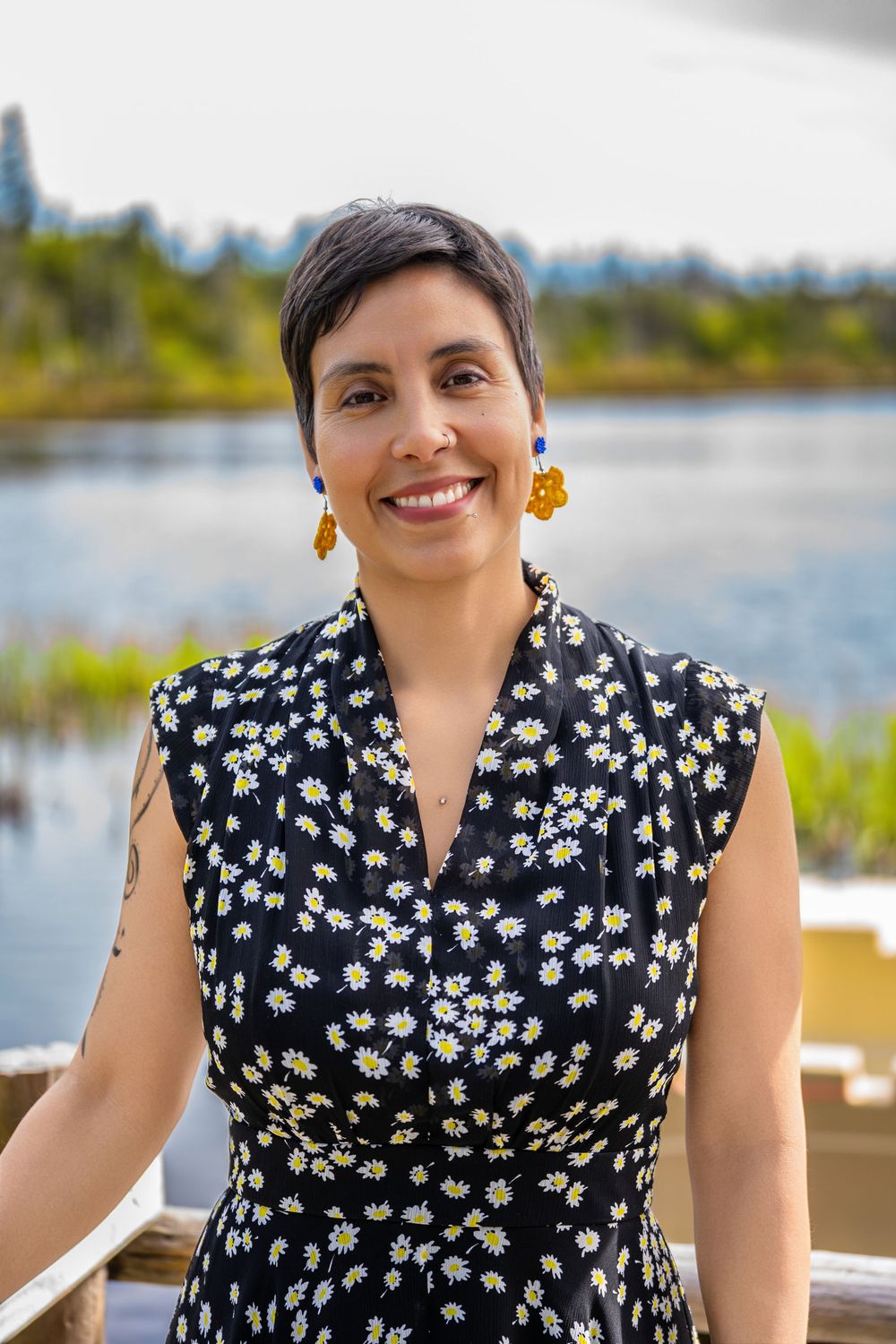 Smiling woman with short hair wearing a floral dress and yellow earrings, standing outdoors near a serene lake.