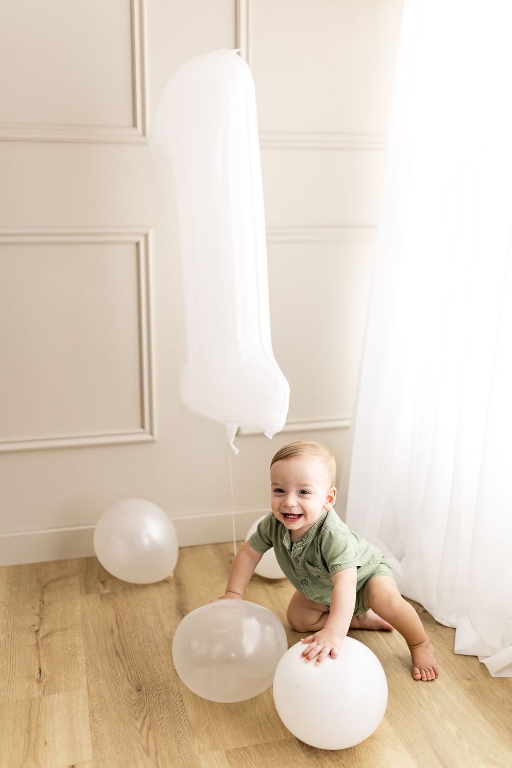 Baby in green onesie plays with white balloons on hardwood floor against white paneled wall.