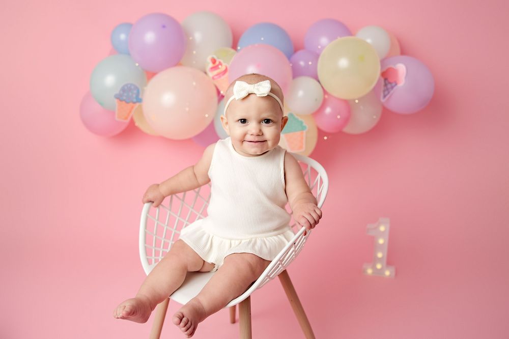 A baby wearing a white dress sits among pastel balloons against a pink backdrop for a first birthday photoshoot.