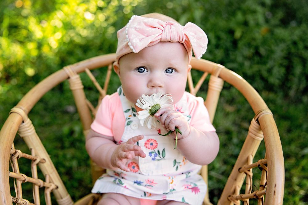 Baby sitting in wicker chair wearing pink outfit and floral headband.