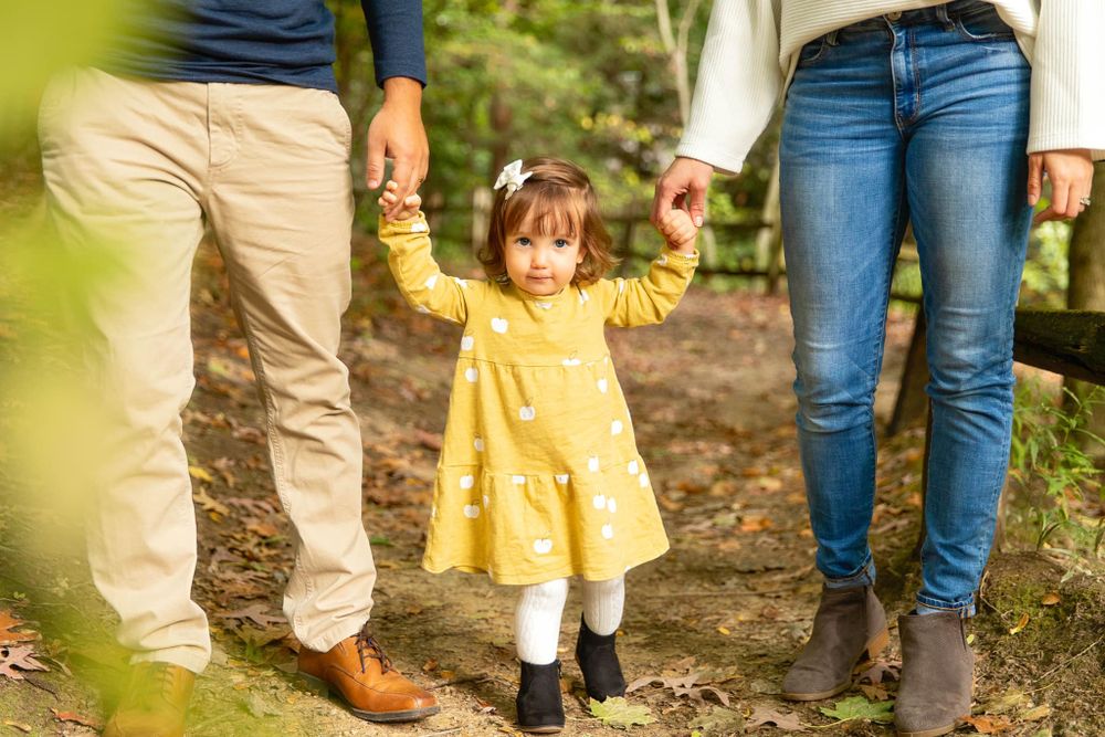 A small child in a bright yellow dress holds hands with their parents while walking on a forest trail.