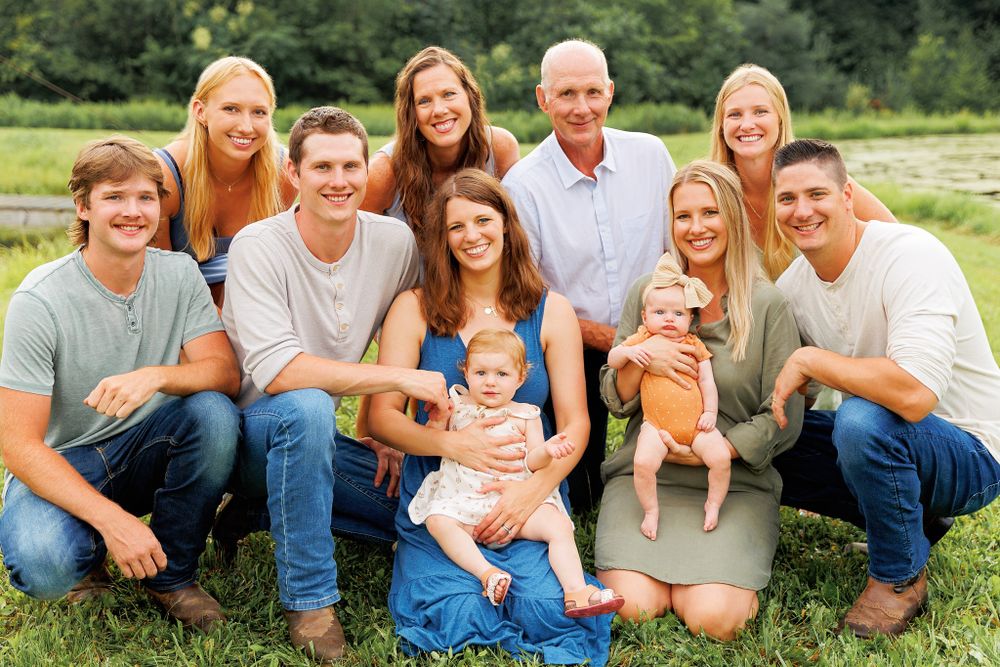 Family group photo in casual attire sitting together outdoors in a green field on a sunny day.