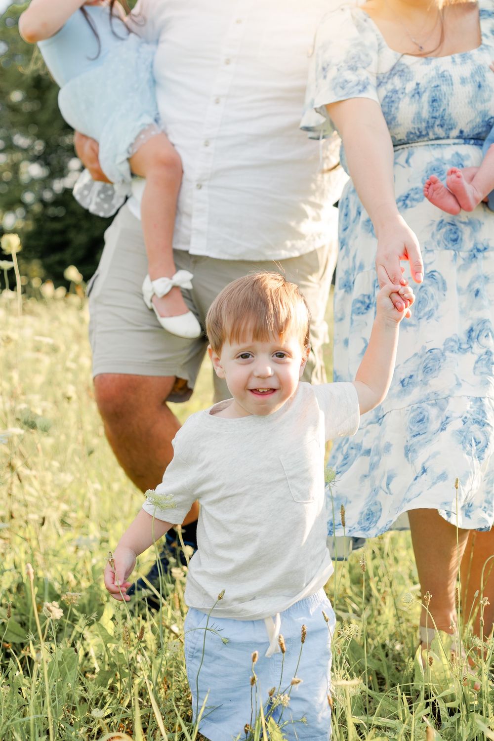 Small child in a white shirt and jeans holding hands with adults in a sunny meadow.