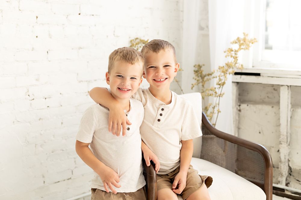 Two young children in white shirts sitting together on a bench against a white brick wall.
