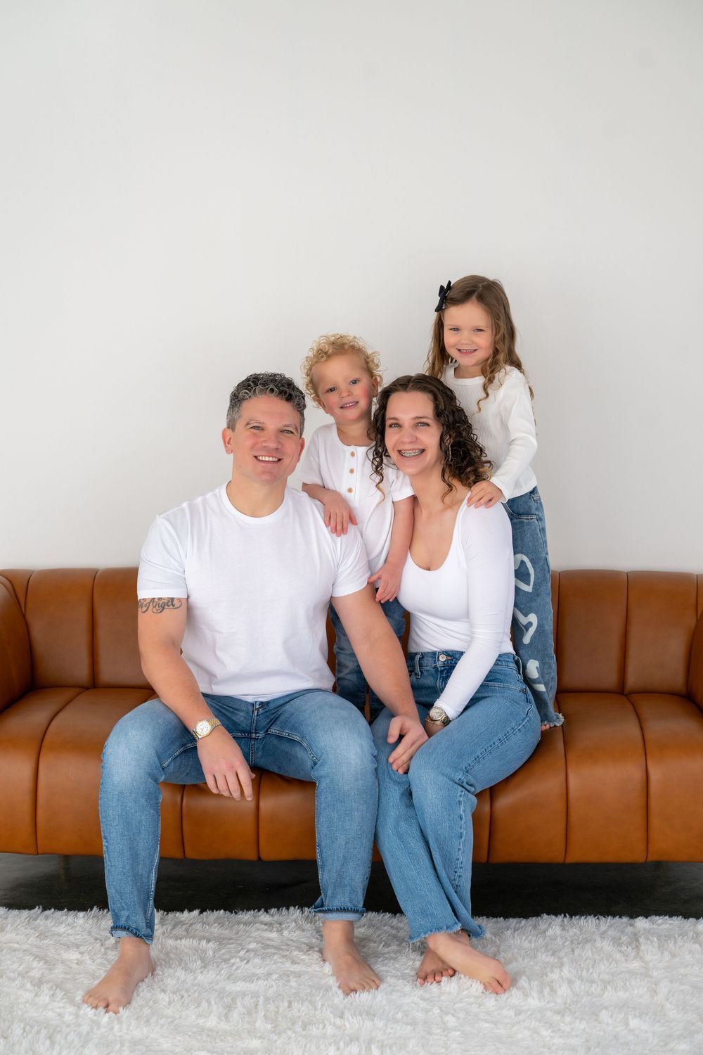 Family wearing matching white t-shirts and jeans sits together on brown leather couch for casual portrait session.