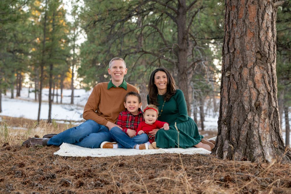 Family of four sitting together on blanket in snowy winter forest setting.