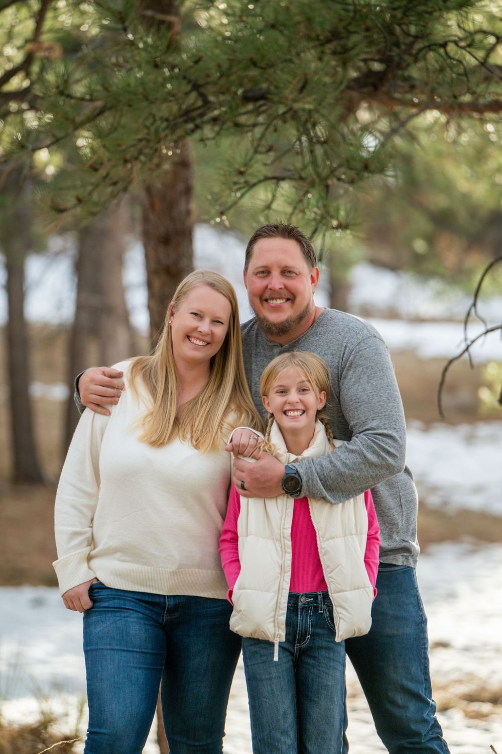 Family of three poses together in winter forest setting wearing casual clothing.