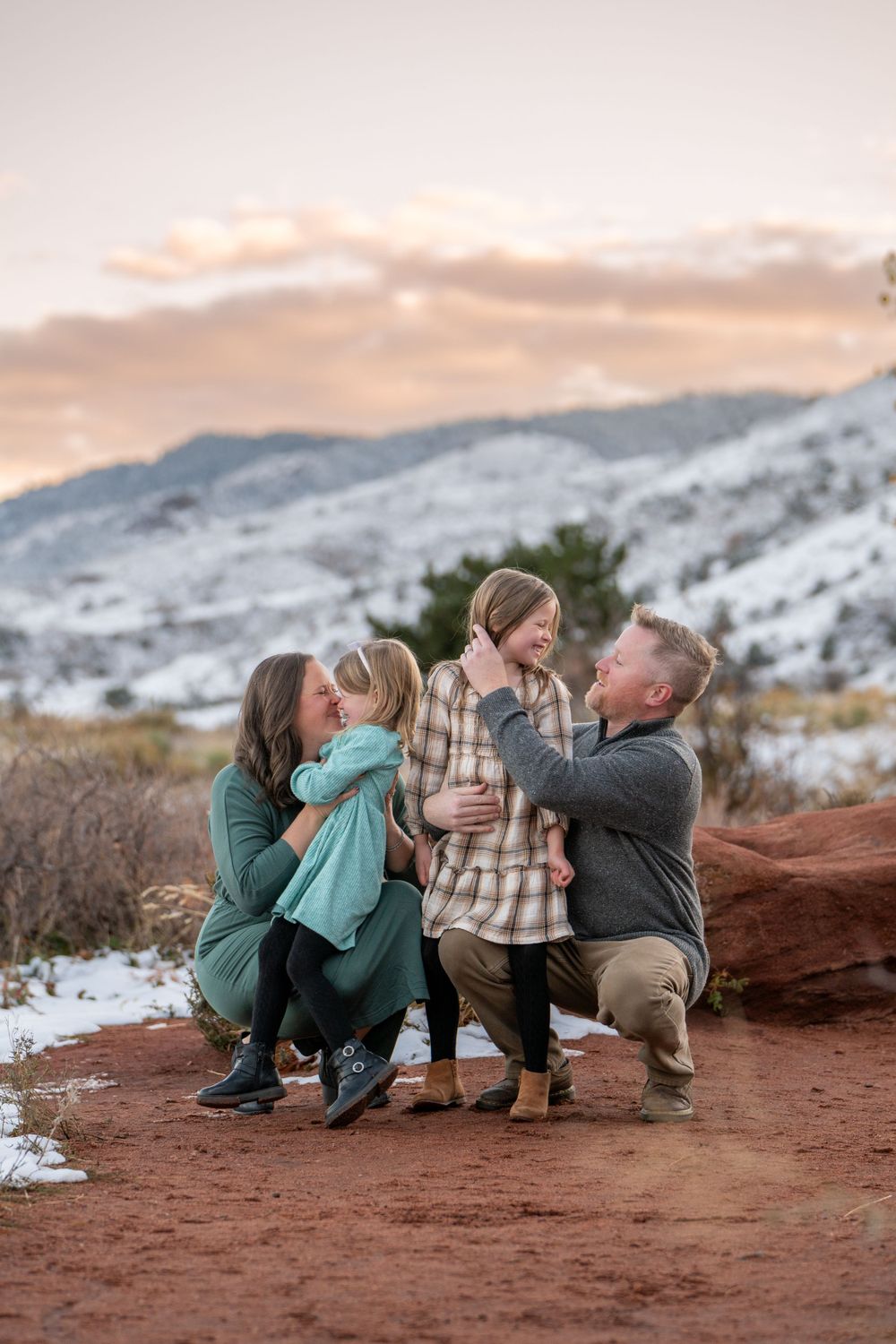 Family sharing intimate moment together on red rocks with snowy mountains in background.