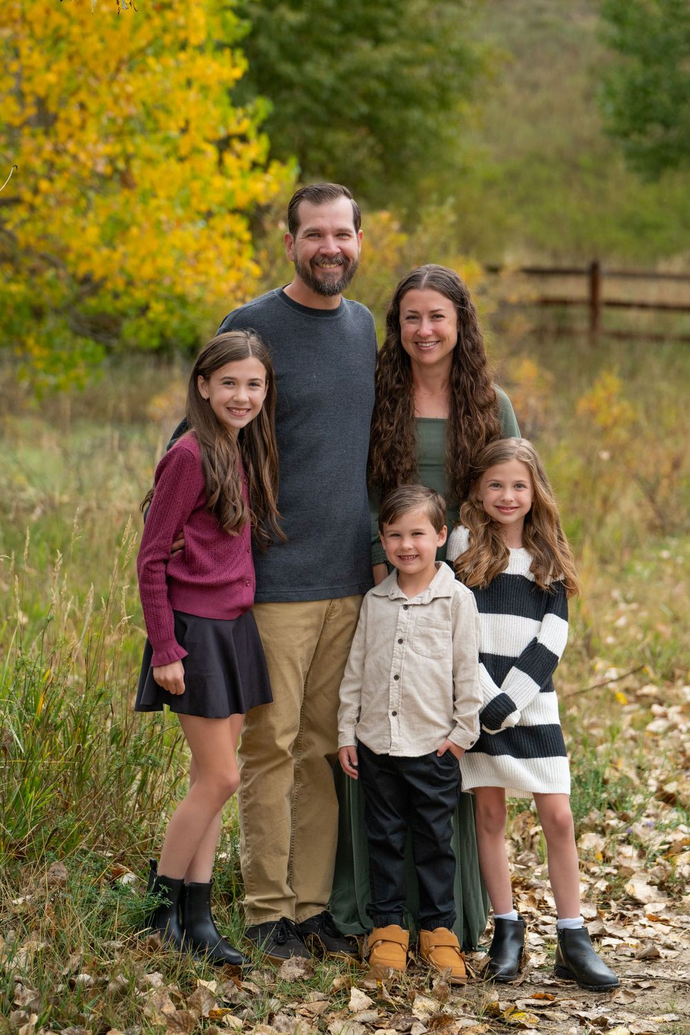 A family poses together outdoors during autumn with yellow foliage and fallen leaves in the background.