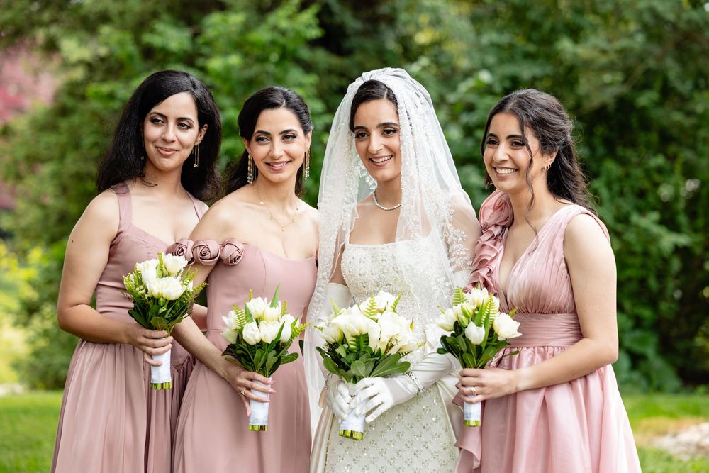 Bride in white lace gown and veil poses with three bridesmaids in blush pink dresses holding white tulip bouquets outdoors.