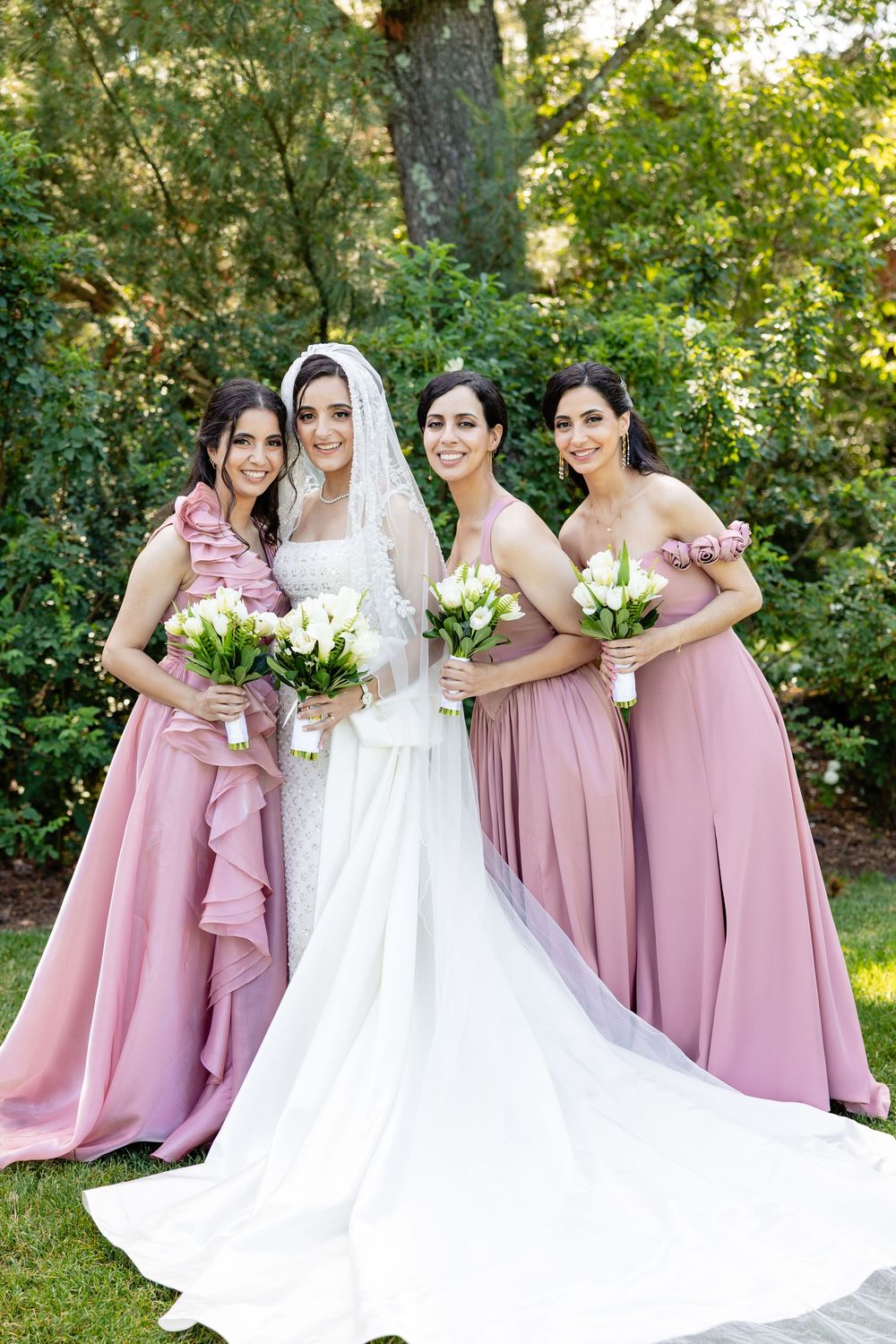 Bride in white gown and veil poses with three bridesmaids in pink dresses holding white bouquets outdoors.