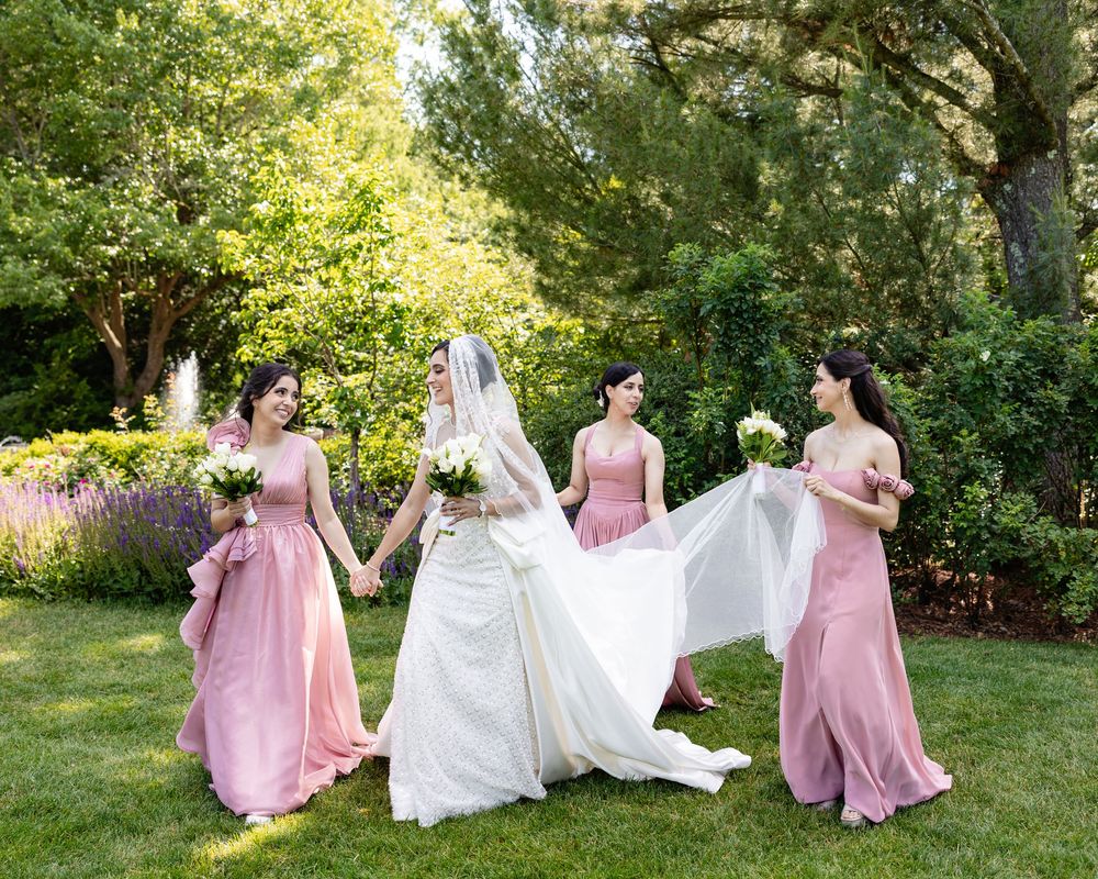Bride in white gown with veil held by three bridesmaids in pink dresses, posing outdoors in a lush garden.