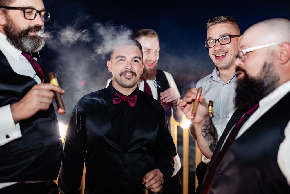 Groom and groomsmen in tuxedos celebrating with cigars and sparklers at a wedding reception.