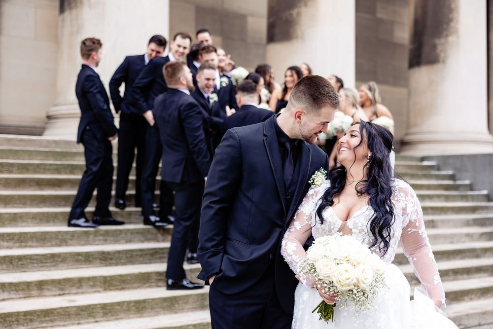 Bride and groom share a romantic kiss on stone steps surrounded by wedding party in black suits.