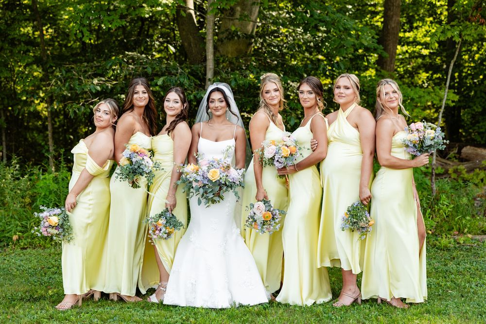 Bride in white gown poses with six bridesmaids in yellow dresses holding bouquets outdoors.